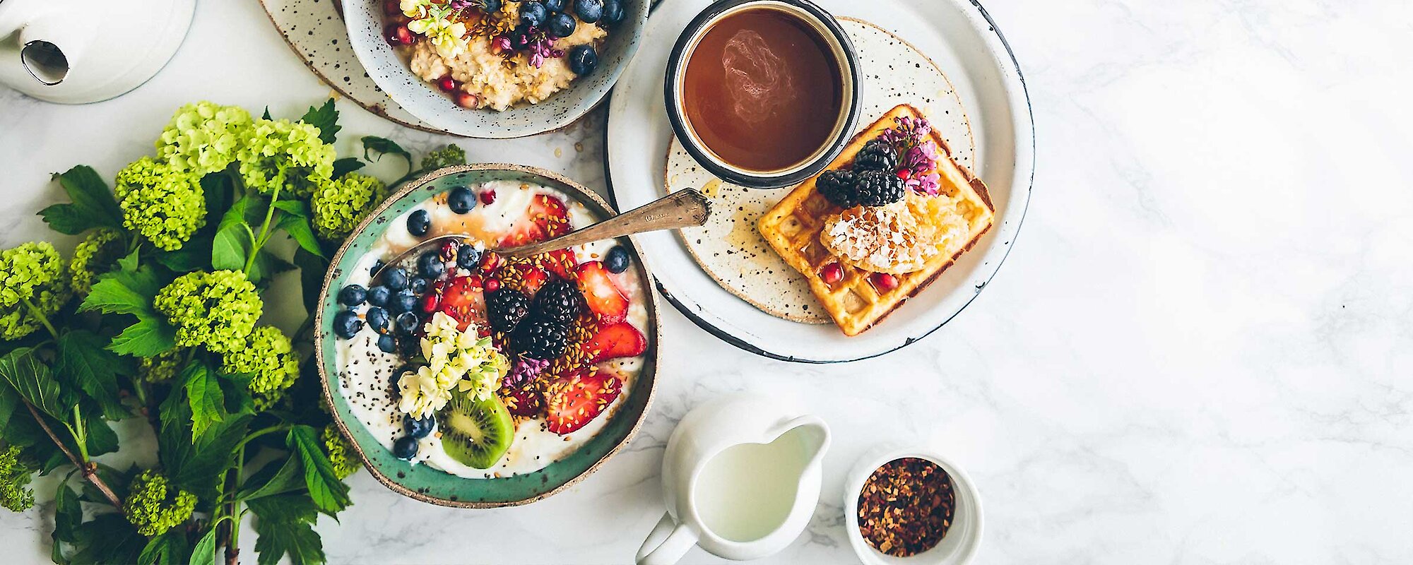 A breakfast spread of porridge, fruit, waffles and coffee lay on a marble table accented with green flowers